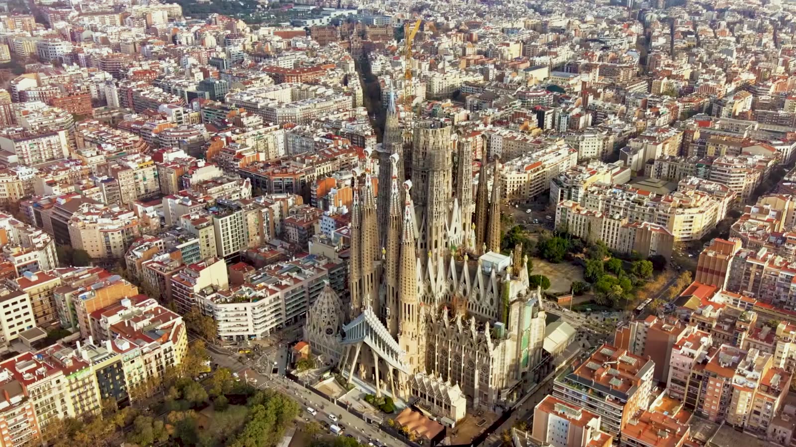 Aerial view of Barcelona's Eixample grid with the Sagrada Família basilica rising from the city center, surrounded by uniform city blocks stretching to the horizon