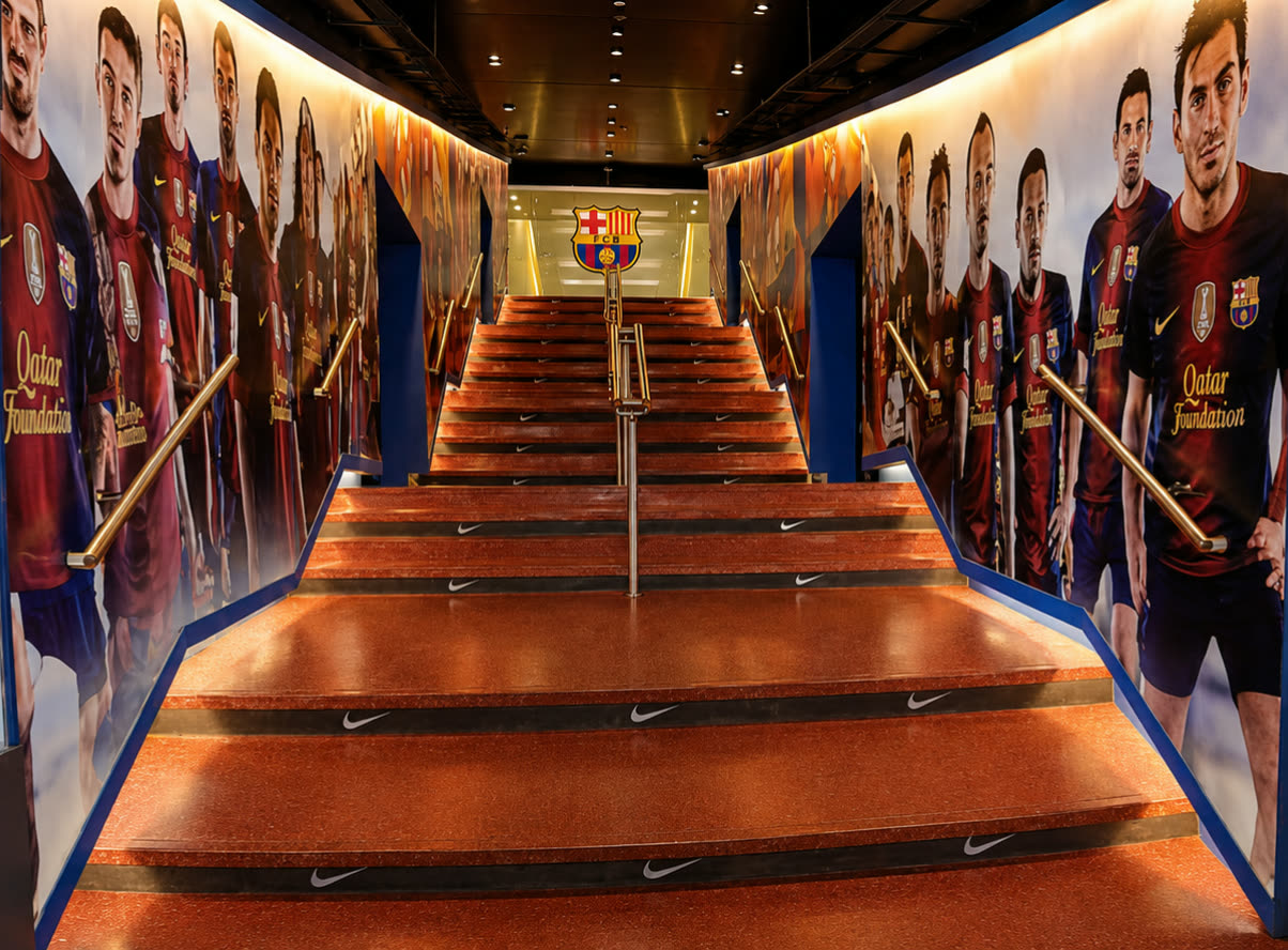The wide stairway inside the Camp Nou museum corridor, walls covered with life-size player imagery and Qatar Foundation sponsorship, FC Barcelona crest at the top of the climb