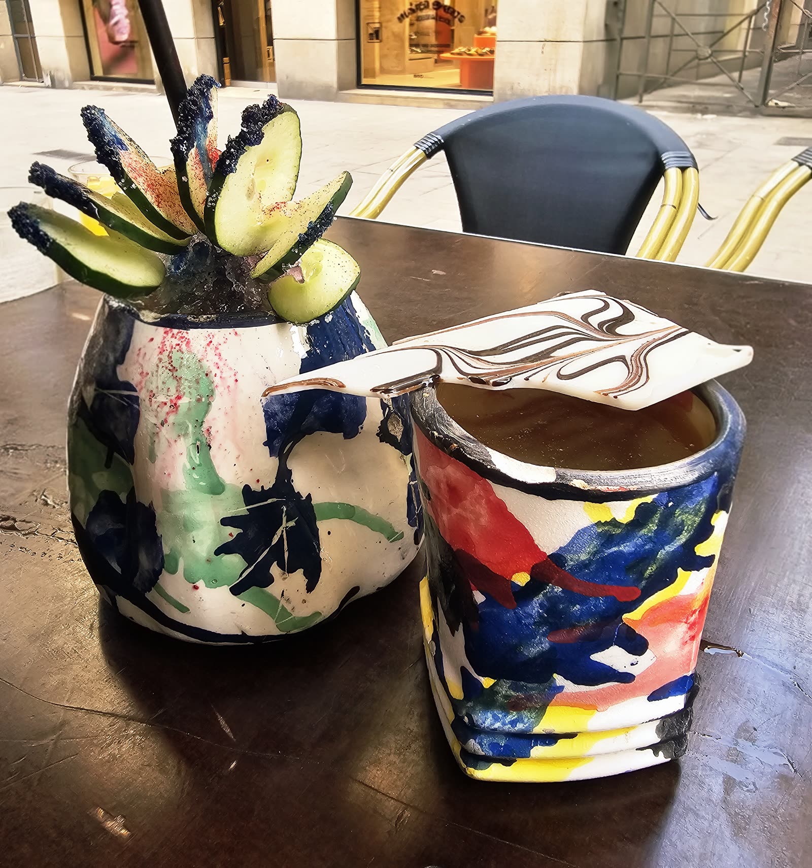 Two hand-painted ceramic cups on a wooden cafe table outside Mariposa Negra — one topped with cucumber slices and a salted rim, the other with a marbled ceramic shield laid across the top, both painted in primary-color brushstrokes