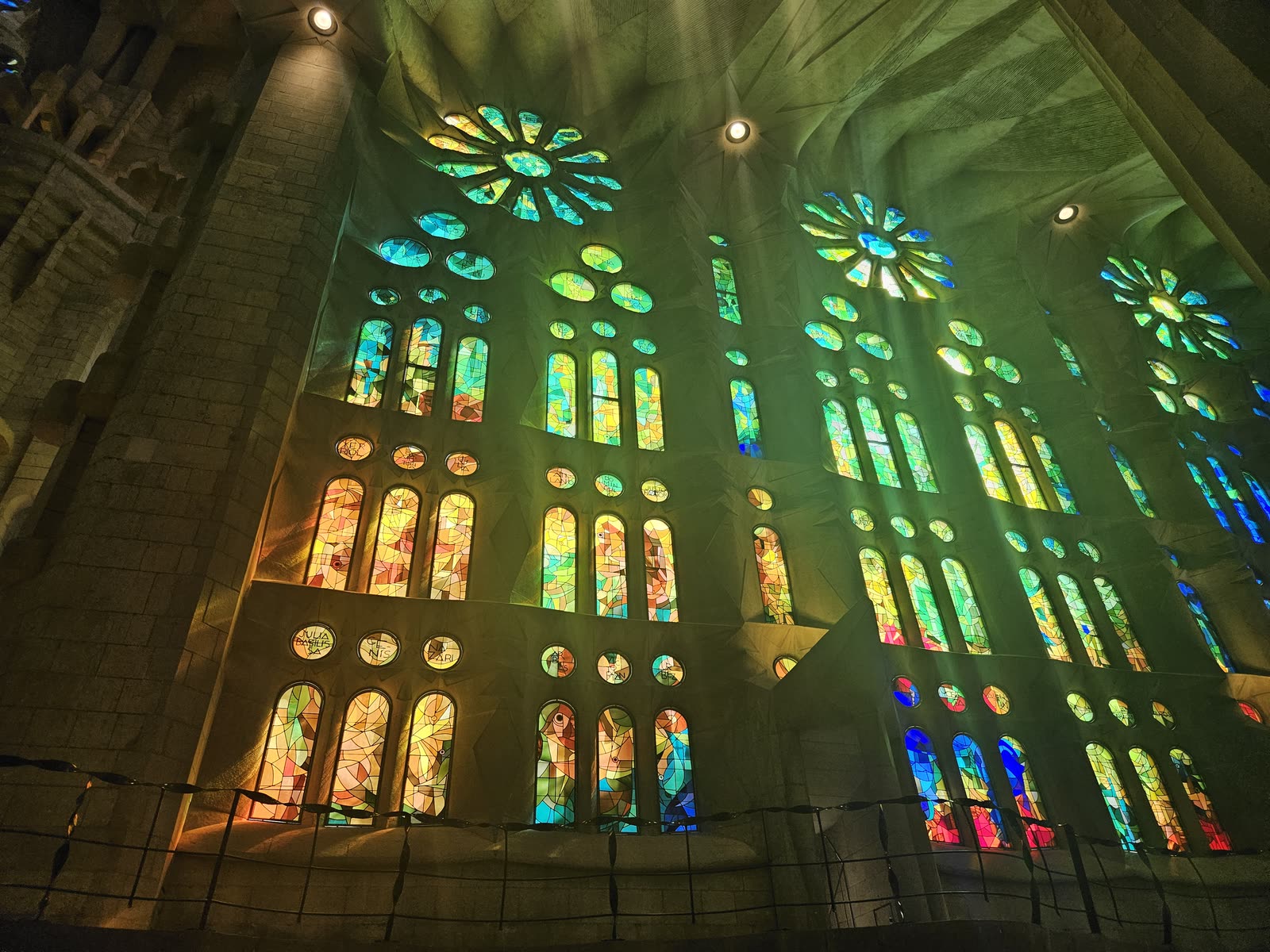 Sagrada Família interior — green and yellow stained-glass light streaming through three rose windows and arched panels, painting the limestone walls in cool jewel tones