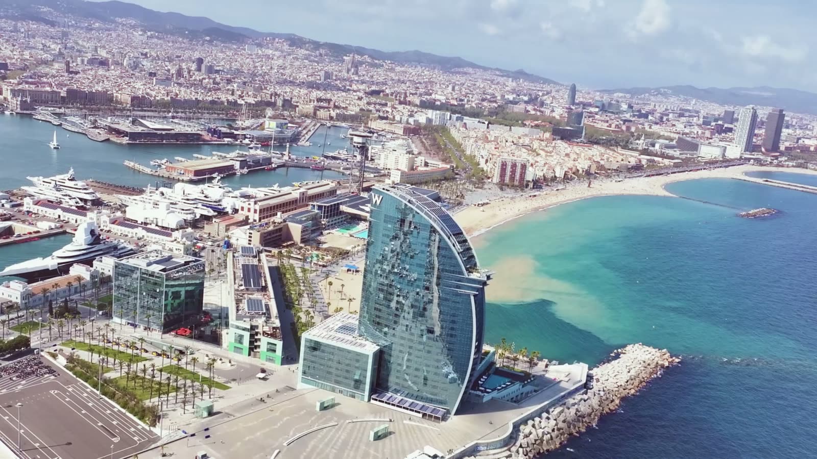 Aerial view of Barcelona's waterfront — the sail-shaped W Hotel rising over Barceloneta beach, the marina to the left, and the city stretching inland to the right