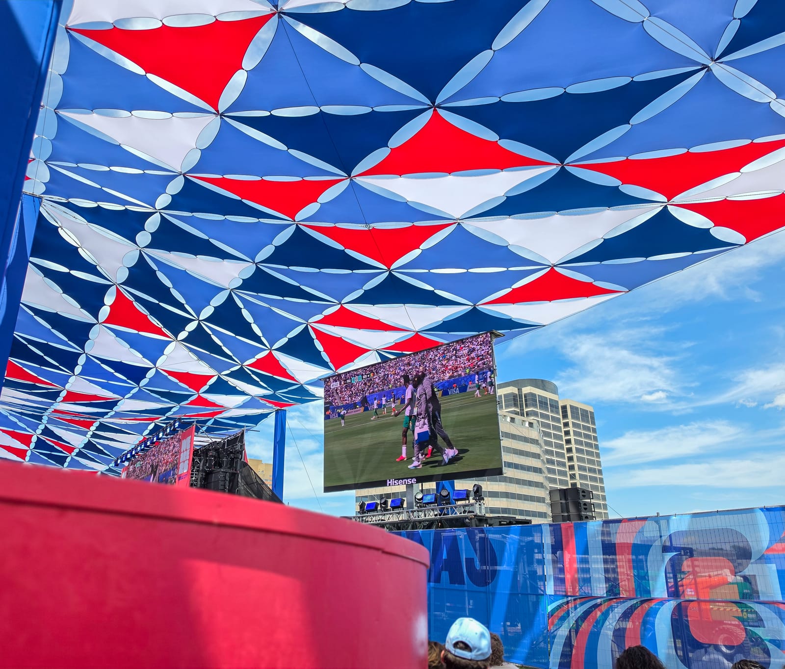 Fans watching a World Cup match on a large screen from under a red, white, and blue shade canopy at the Kansas City Fan Festival