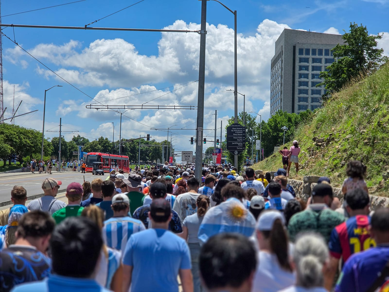 World Cup fans in Argentina blue and white walking along a Kansas City road toward the venue under a bright summer sky, a transit bus ahead