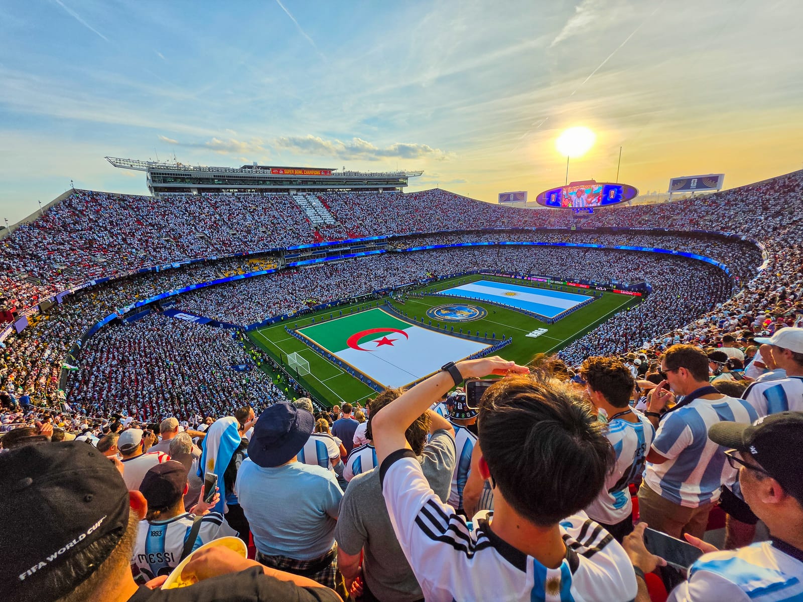 Packed Kansas City Stadium at golden hour during the World Cup, with large Argentina and Algeria flags spread across the pitch before kickoff.