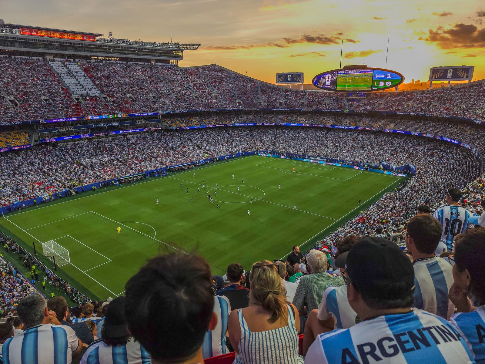 View from the stands of a World Cup match in play at sunset in Kansas City, with fans in Argentina shirts in the foreground