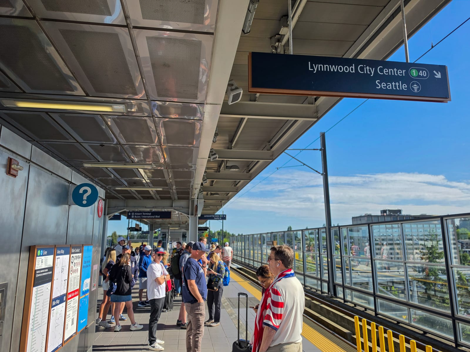 World Cup fans in United States colors waiting on a Sound Transit Link light rail platform on matchday, under a Lynnwood City Center and Seattle direction sign
