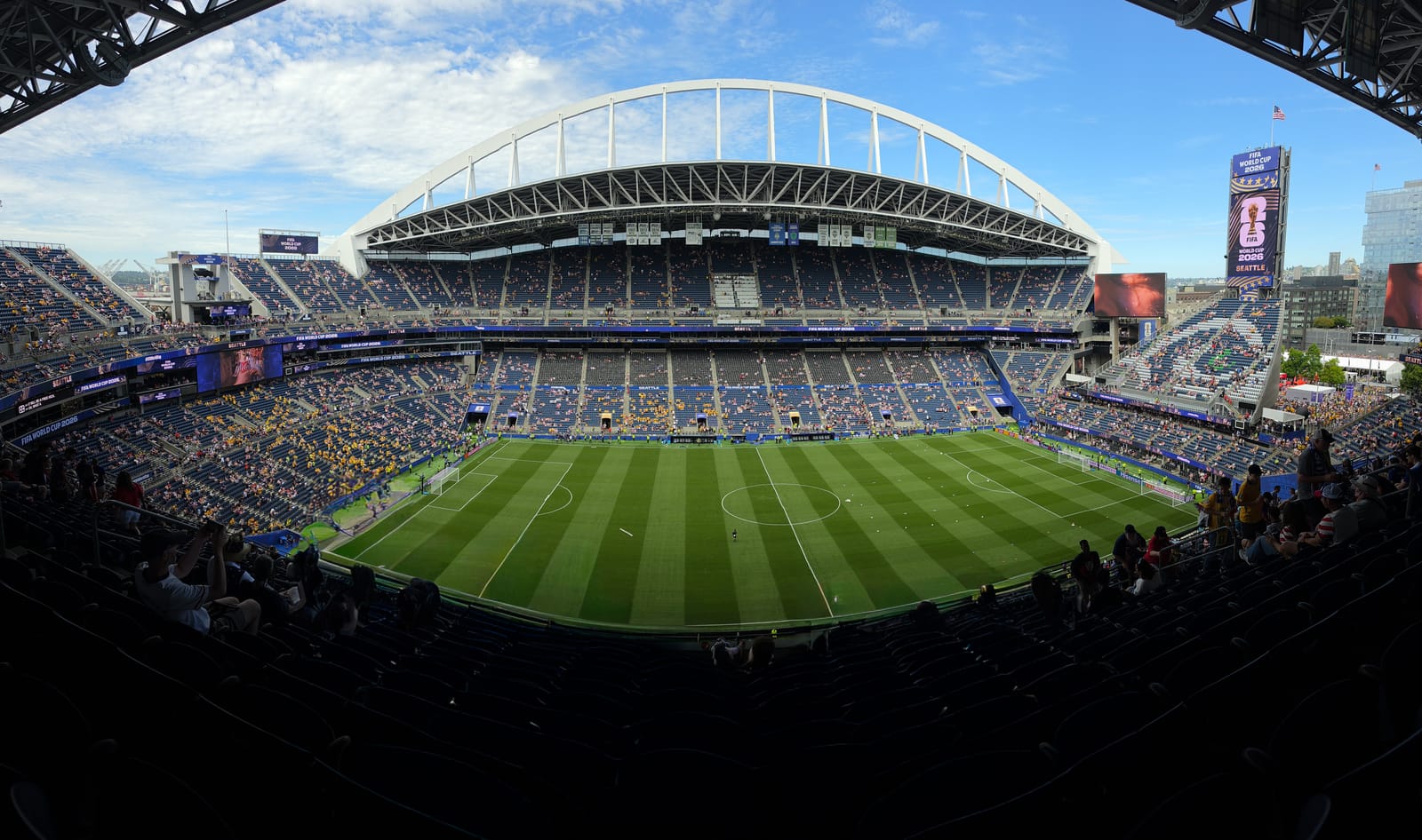 View from shaded seats inside the stadium before a World Cup match in Seattle, looking across the green pitch to the open end of the stadium and the skyline beyond